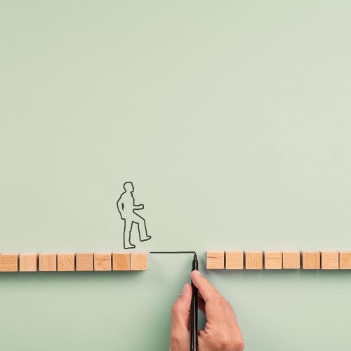 Male hand drawing a bridge between two rows of wooden blocks for a silhouetted man to walk across. Over blue background with plenty of copy space.
